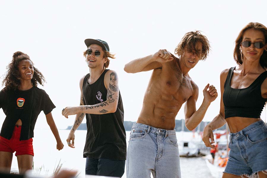 Imagen de dos hombres y dos mujer que parecen bailando y de fondo se ve playa y botes un poco borrosos